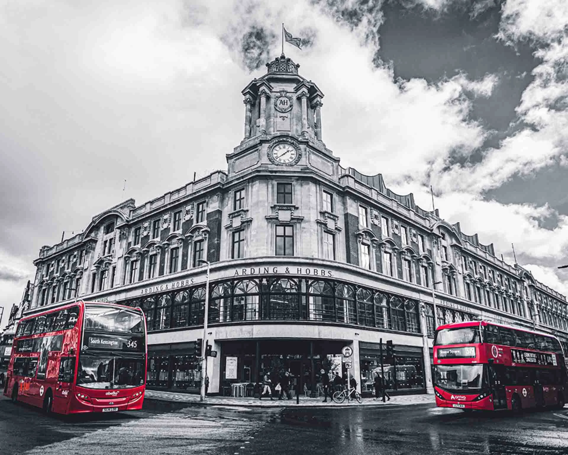 Complete black and white photo except for two double-decker London buses in the bottom left and right of the frame. Shot in a street photography style there is an old Arcadian building in the centre of the frame and two busses parked at an intersection to the left and right of that building